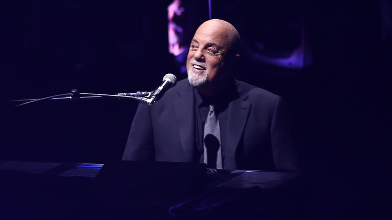Billy Joel smiling sitting behind microphone and piano