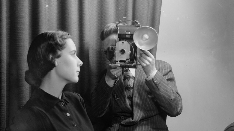 A polaroid instant camera in 1949 in a black and white photo of a man taking a photoof a woman