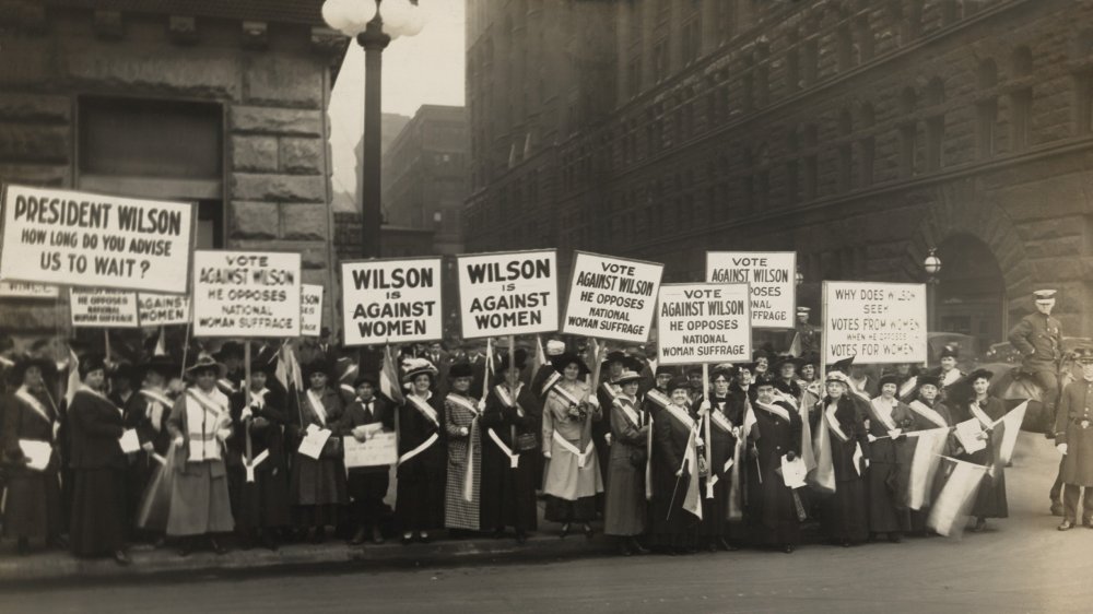 A crowd of protesting suffragettes in 1916 carrying signs saying "Wilson is against women."