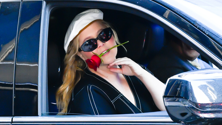 Amy Schumer in sunglasses looking out car window with rose in mouth