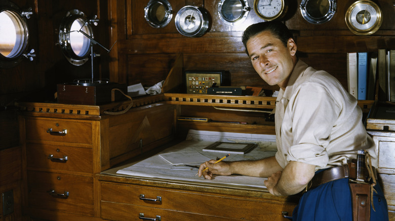 Errol Flynn in white shirt standing at desk writing with pencil