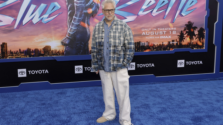 James Gunn in plaid and khakis in fromt of Blue Beetle poster