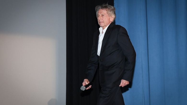 Roman Polanski in black suit holding microphone in front of blue curtain