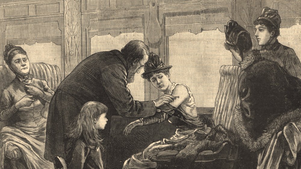 A doctor leans over and pricks the arm of a seated woman in a train car. Several other people either stand or sit and watch.