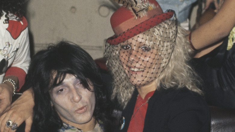 Sable Starr smiling and sitting on a male rocker's lap in a red hat and black veil