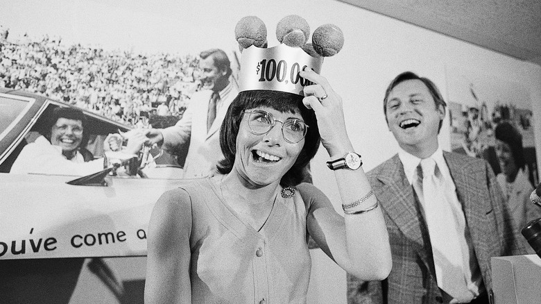 Billie Jean King wearing a crown adorned with tennis balls and a $`100,000 figure