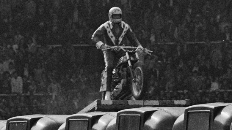 Evel Knievel riding motorbike up ramp over cars
