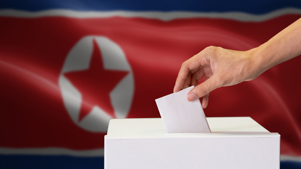 A hand placing a ballot into a white box before a North Korean flag